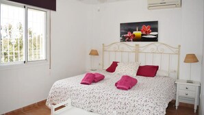 A bedroom with a bed, two bedside tables, a ceiling fan, an air conditioning unit, and a window with a view of greenery. - Villa Buenavista in C Mpeta (Torrox)
