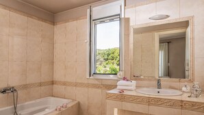 A bathroom with a bathtub, a sink, a mirror, and a window with a view of greenery. - Villa Chloe in Rethymno (Rethymno)