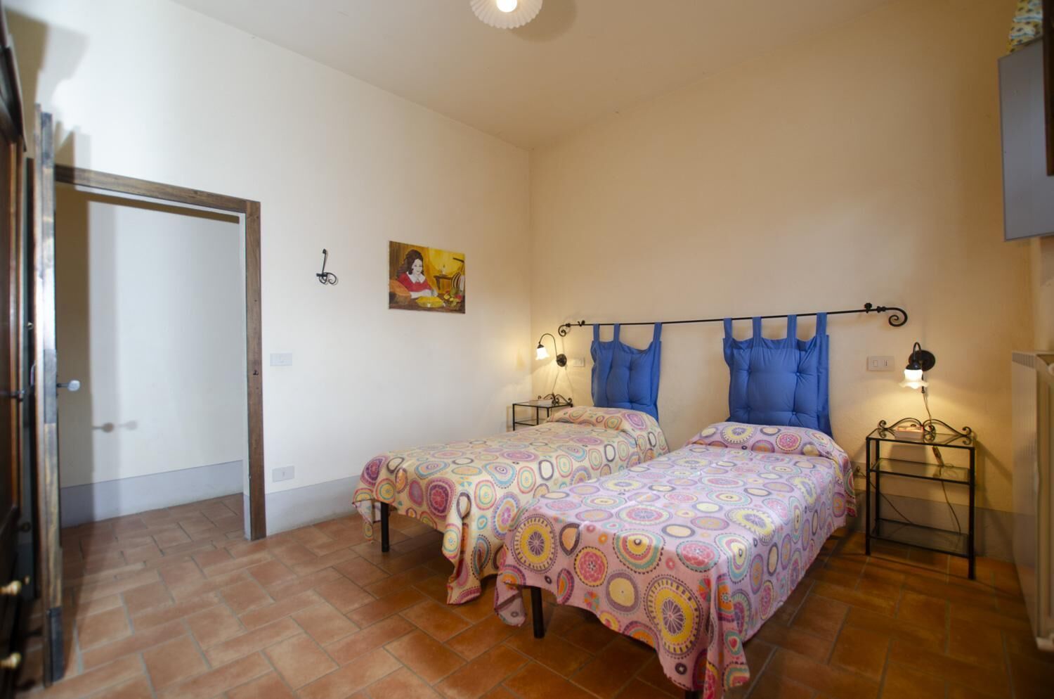 A room with two beds, a door, a wall-mounted picture, and a small table with a lamp.