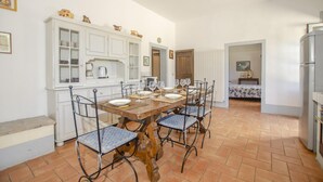 A dining area with a wooden table set for four, surrounded by chairs with blue cushions. A white cabinet with glass doors is to the left, and a refrigerator is visible to the right. - Casale del Conte in Pienza (Sarteano)