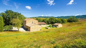 A stone building with a tiled roof, surrounded by a grassy field and trees, with hills in the background. - Casale del Conte in Pienza (Sarteano)