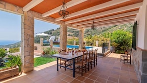 Un espacio de comedor al aire libre con techo y vistas a una piscina y colinas.