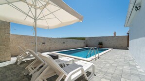 Una piscina con sillas de descanso blancas y una sombrilla, rodeada de muros de piedra y un patio empedrado.