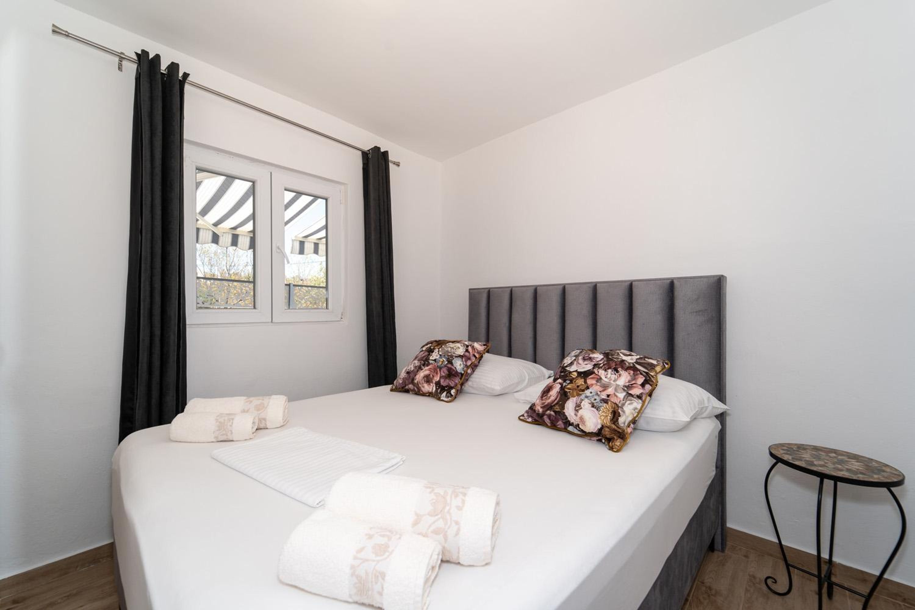 Een keurig opgemaakt bed met bloemenkussens, een klein rond tafeltje en een raam met gordijnen.