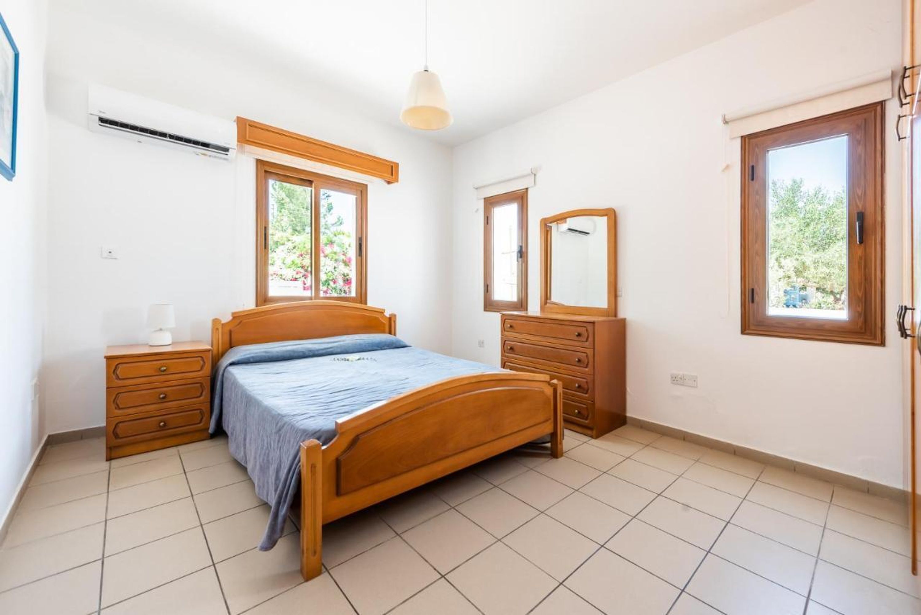 A bedroom with a wooden bed, a dresser, a mirror, and a window with a view of greenery.