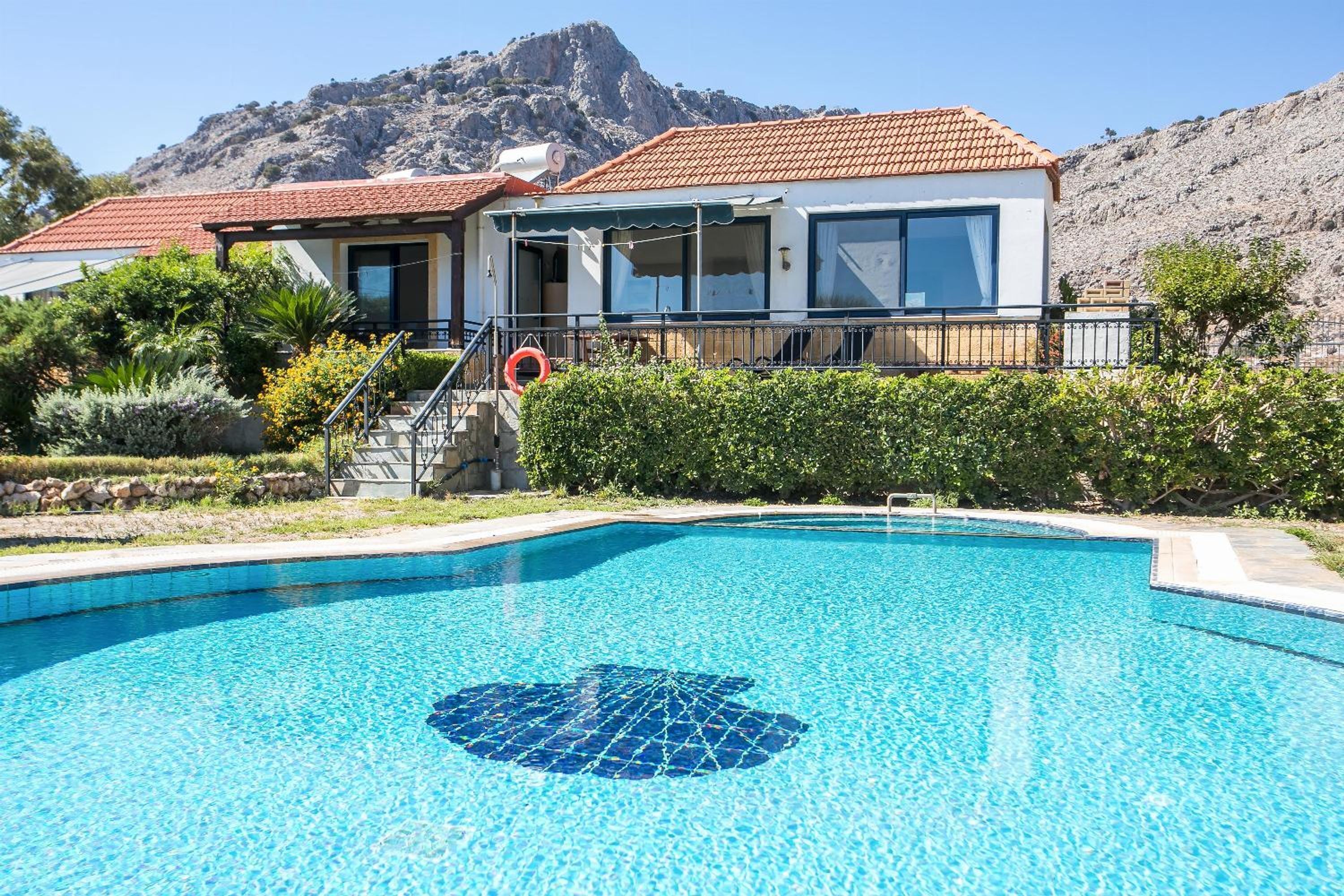 A swimming pool with a tiled design, a house with a red-tiled roof, and a mountainous backdrop.