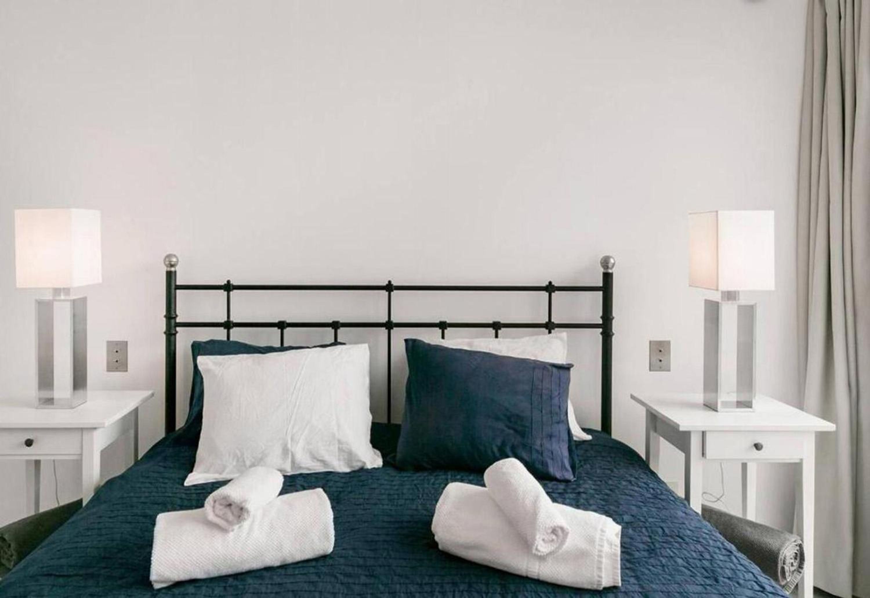 A neatly made bed with white and dark blue pillows, two bedside lamps, and rolled towels on the bed.