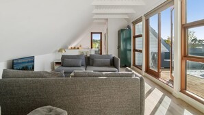 Une chambre de style loft moderne, avec un plafond en pente, de grandes fenêtres, un canapé gris et une vue sur l’extérieur.