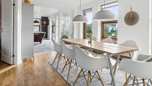 Un espace de repas moderne avec une table en bois et des chaises blanches, de grandes suspensions et une vue sur l’extérieur.