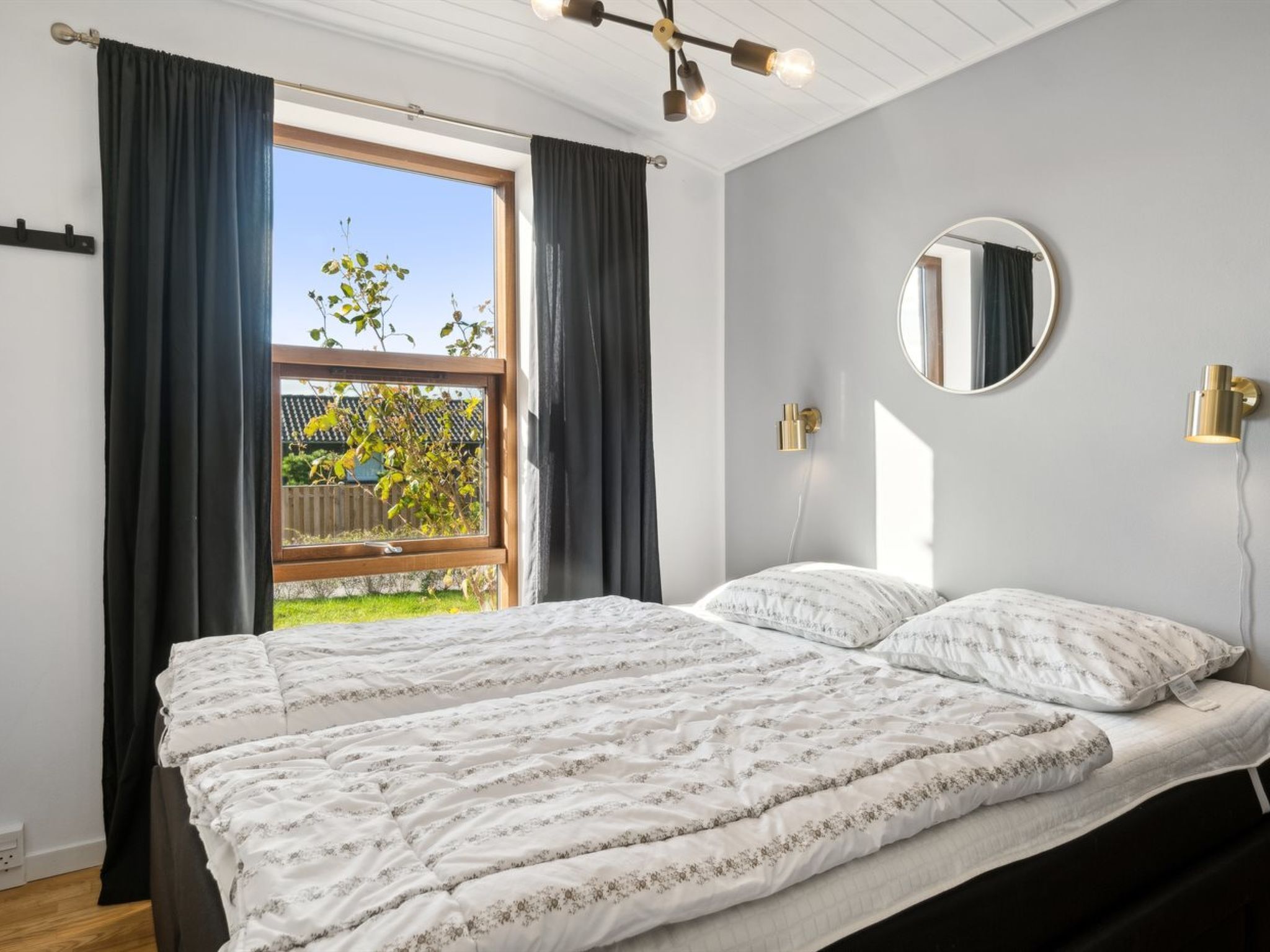 A bedroom with a large bed, a window with curtains, a round mirror, and a ceiling light fixture.