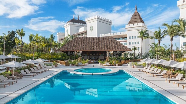 Un espace piscine avec des chaises longues et des parasols, un bar et un bâtiment doté d’un toit et de tours à l’architecture particulière.