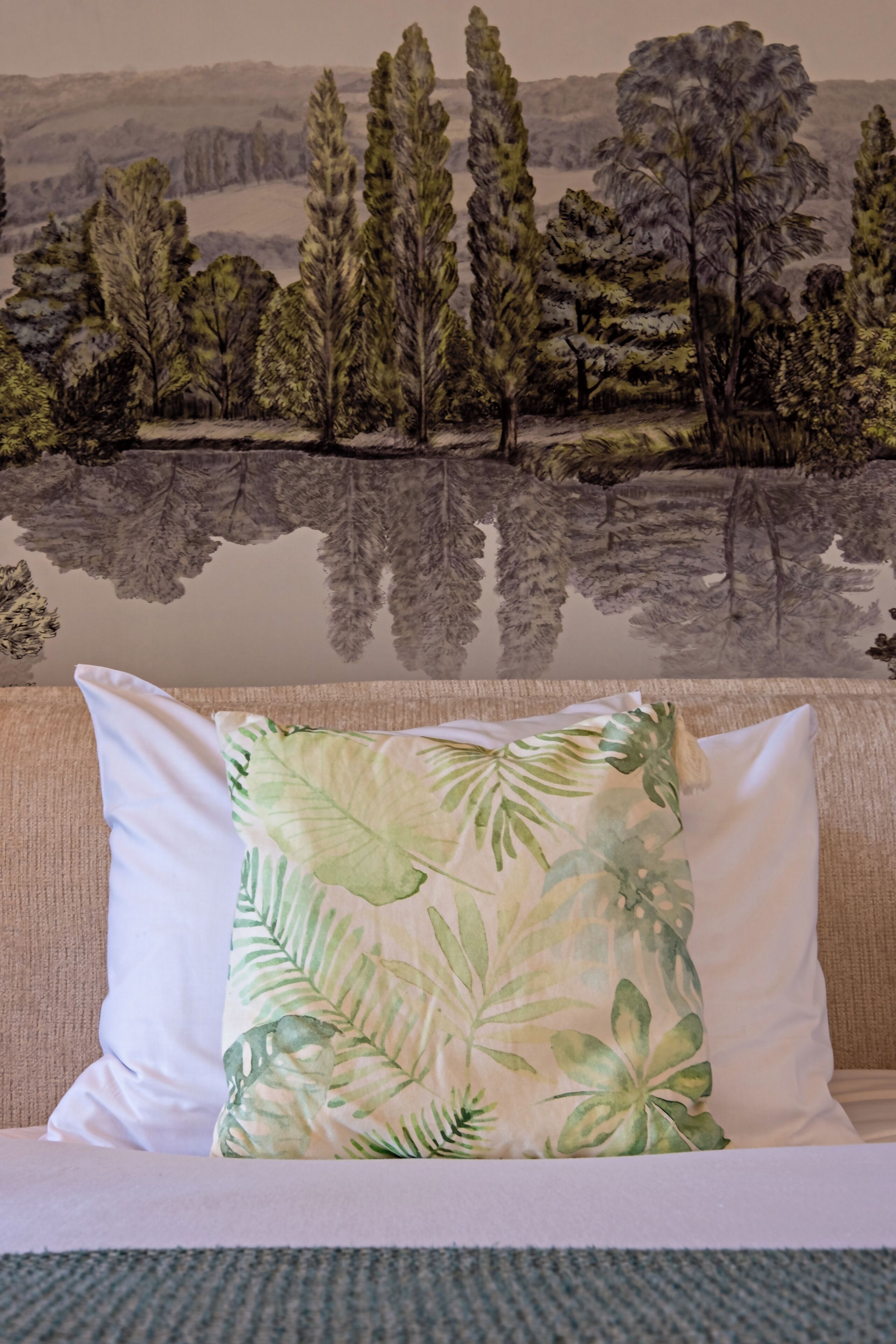 A room with a beige sofa, a white pillow with a tropical leaf pattern, and a mural of a landscape with trees and a body of water.