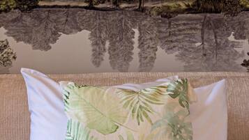 A room with a beige sofa, a white pillow with a tropical leaf pattern, and a mural of a landscape with trees and a body of water.
