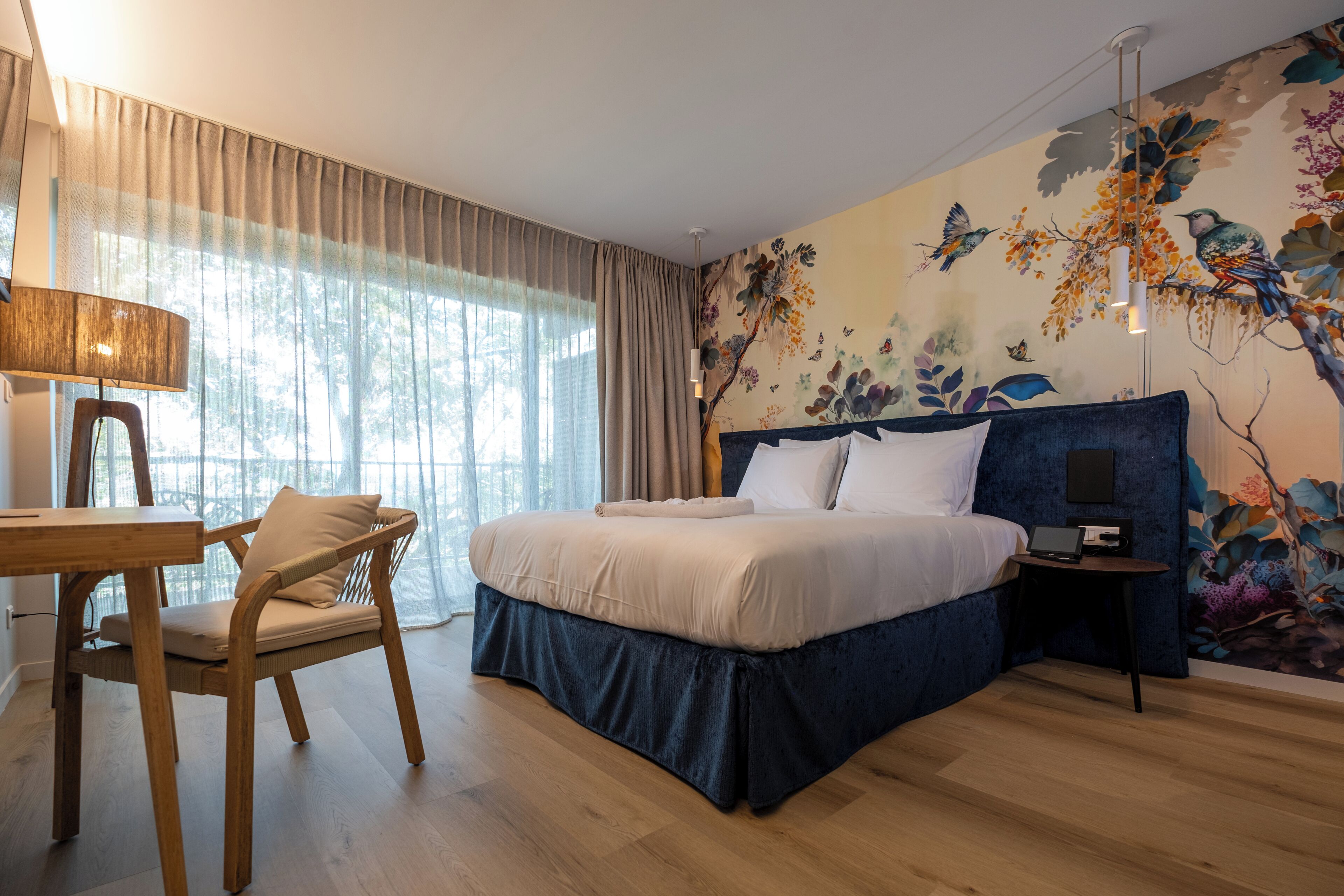 Un dormitorio con cama, escritorio y silla. La habitación tiene un ventanal con cortinas, un mural en la pared y una lámpara colgante.