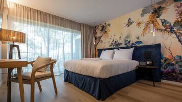 Un dormitorio con cama, escritorio y silla. La habitación tiene un ventanal con cortinas, un mural en la pared y una lámpara colgante.
