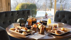 Una mesa de desayuno con diversos alimentos, como pasteles, frutas y bebidas, en un espacio con ventanas amplias y vista a una piscina.