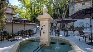 Una fuente de piedra con caños de agua, rodeada de asientos exteriores con sombrillas, con árboles y un edificio de techo a la vista.