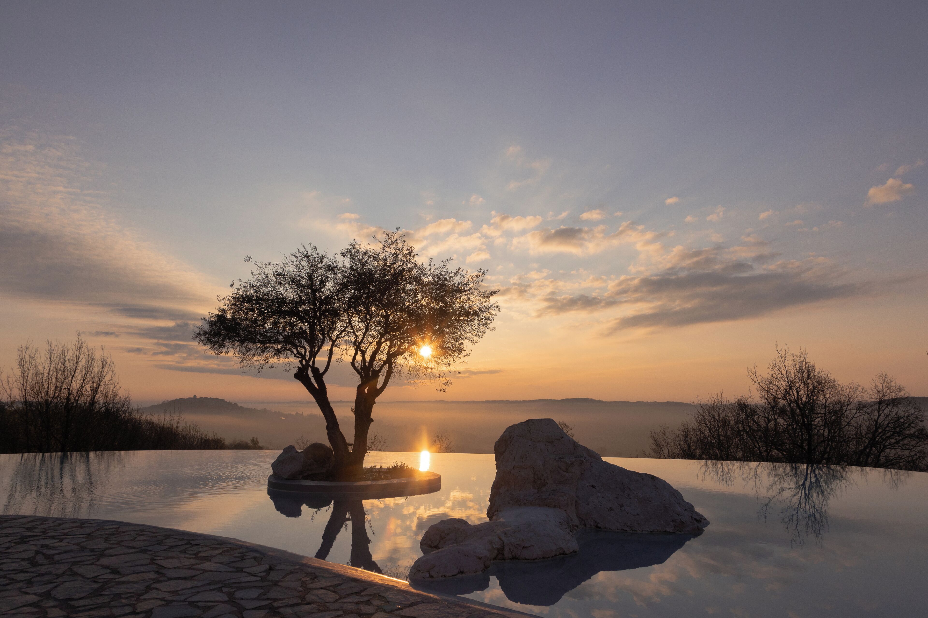 A serene outdoor pool area with a tree and rocks, surrounded by a clear sky and distant hills.
