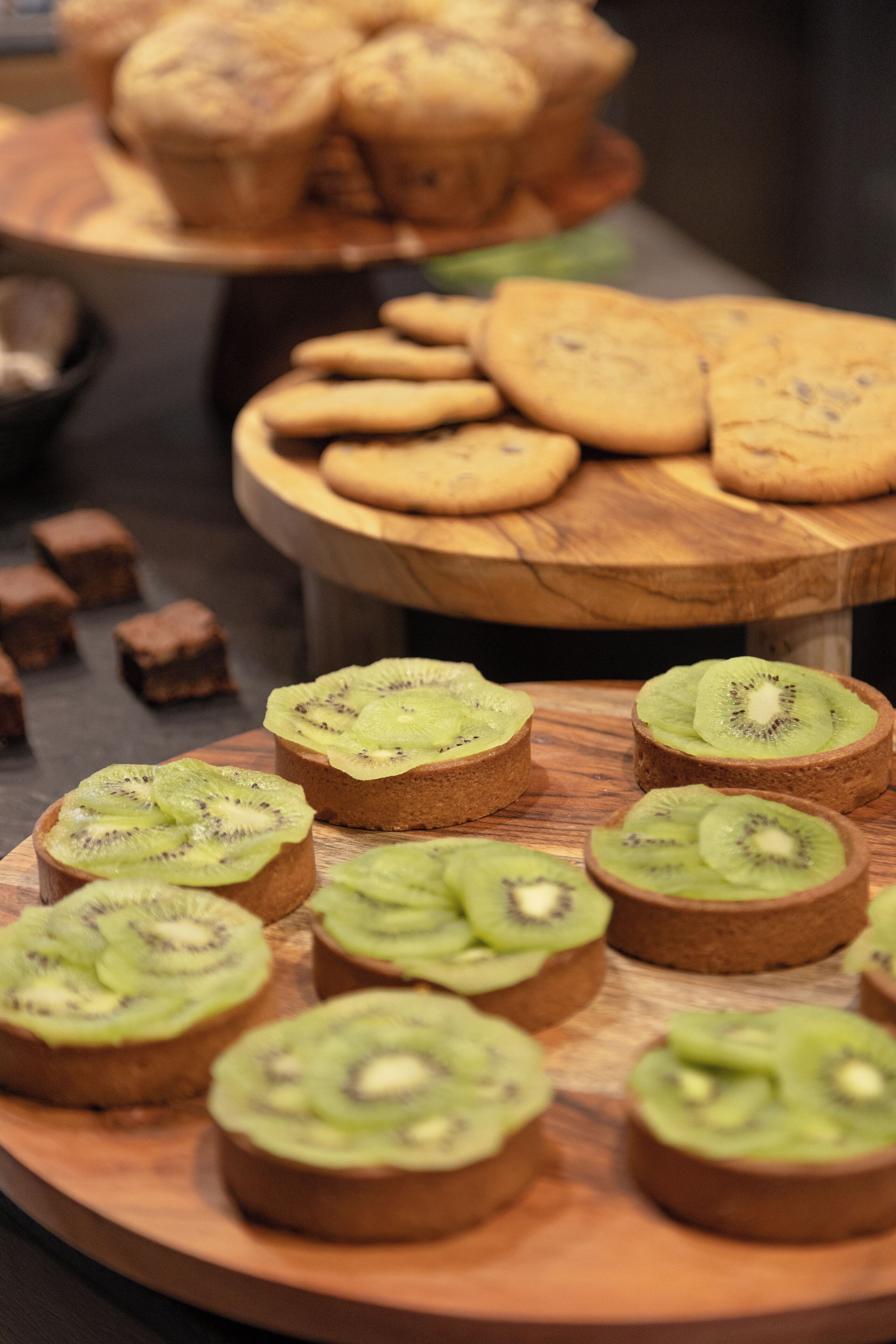 A wooden board with sliced kiwi on top of bread, a wooden board with cookies, and a wooden board with brownies.