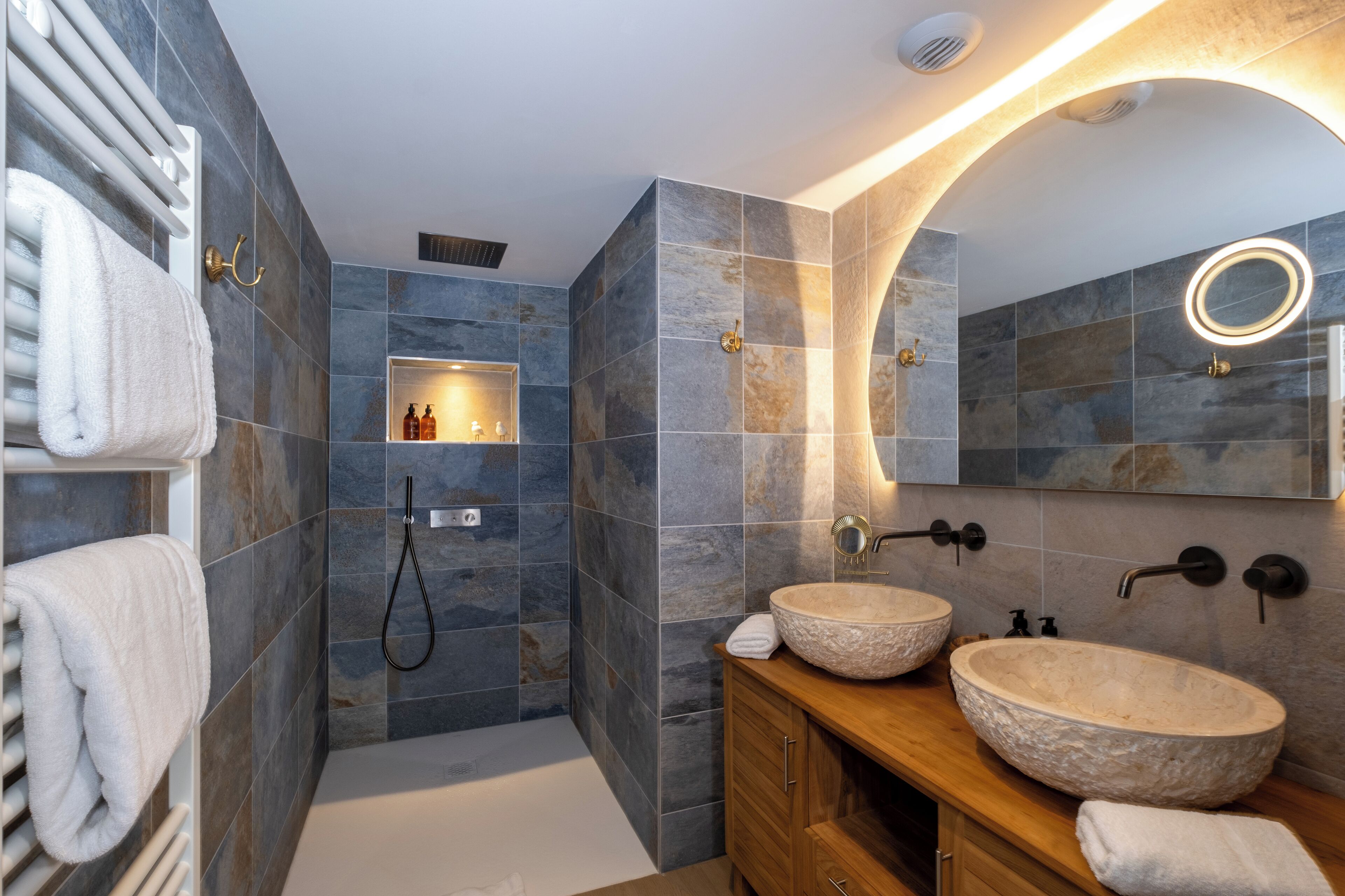 A modern bathroom with stone tiles, a wooden vanity, two large stone sinks, a handheld showerhead, and a built-in shelf with toiletries.