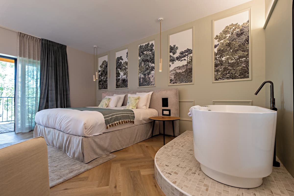 A modern bedroom with a large bed, a round bathtub, and a view of trees through the window.