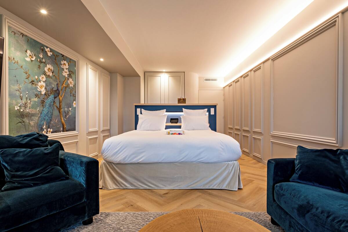A modern bedroom with a large bed, two dark sofas, a wooden coffee table, and a floral painting on the wall.