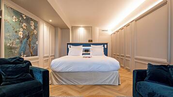 A modern bedroom with a large bed, two dark sofas, a wooden coffee table, and a floral painting on the wall.