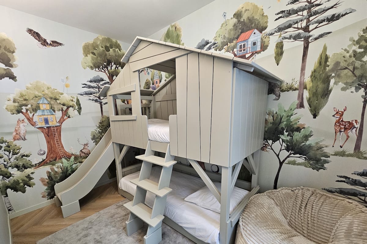 A child's bunk bed with a slide, a round cushioned seating area, and a mural of forest animals and trees on the wall.