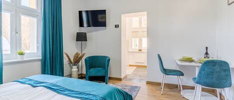 Quarto com cama, escrivaninha, cadeiras, televisão e janela com cortinas.
