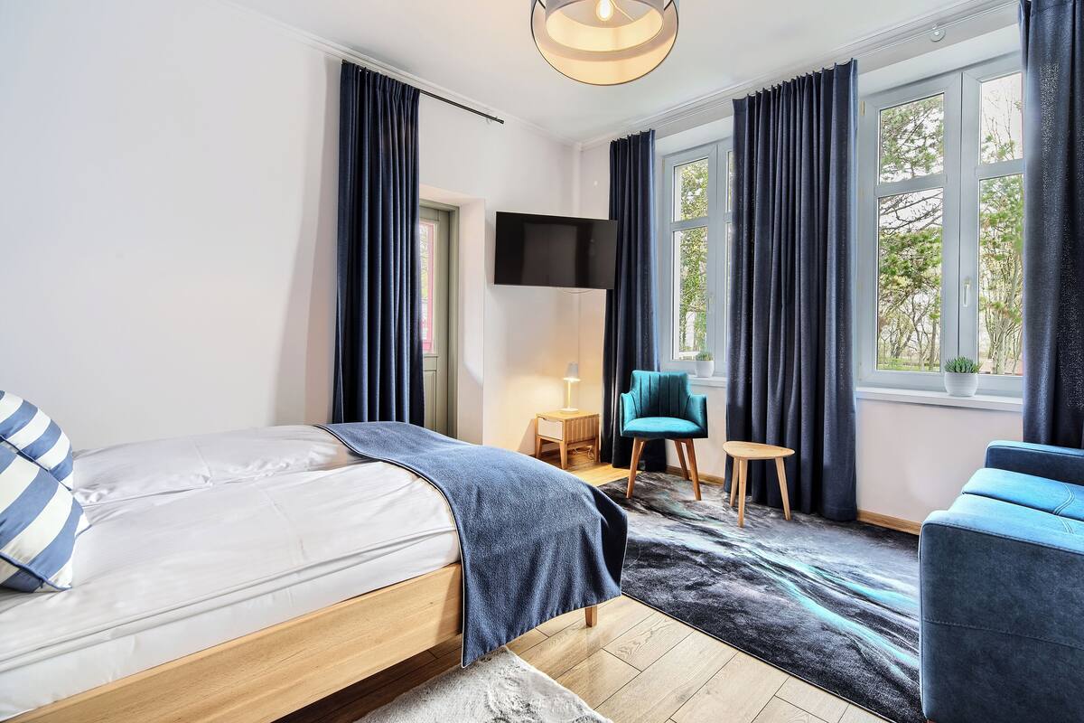Una habitación de hotel moderna con una cama, un sillón azul, una mesita pequeña y un ventanal con cortinas.