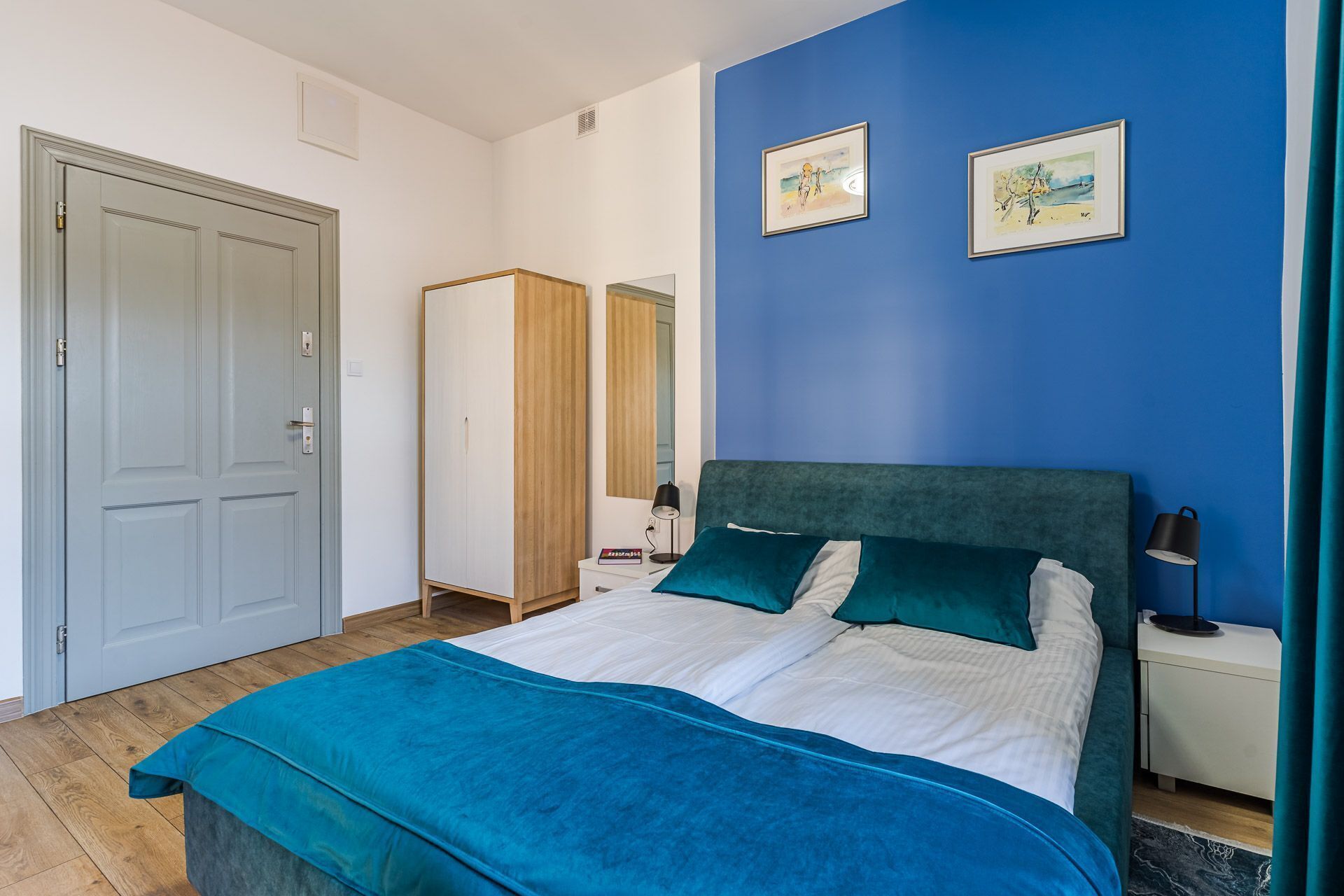 A bedroom with a bed, a nightstand, a wardrobe, and two framed artworks on the wall.