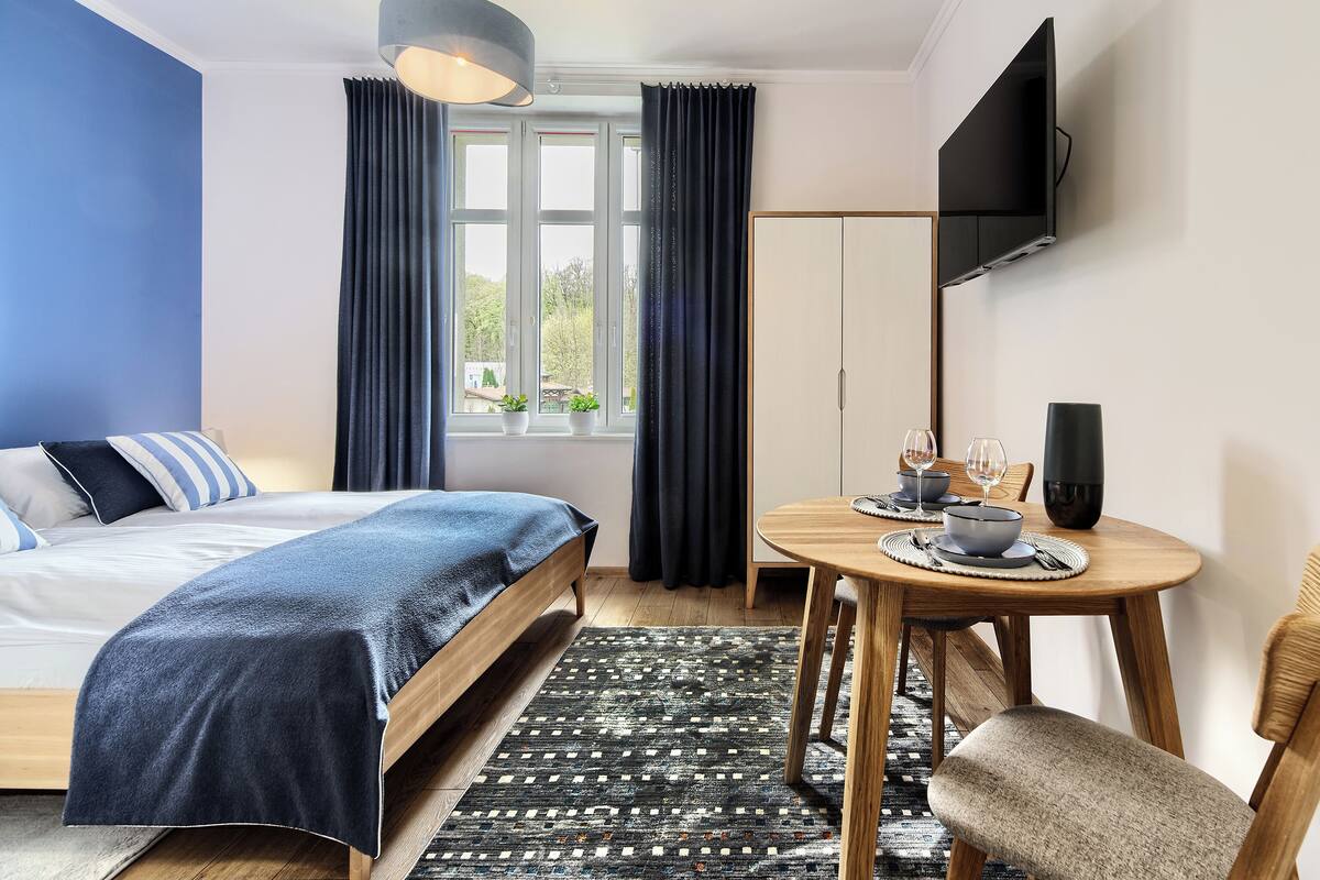Habitación de hotel moderna con una cama, una mesa de comedor con sillas, un televisor montado en la pared y un ventanal con cortinas.