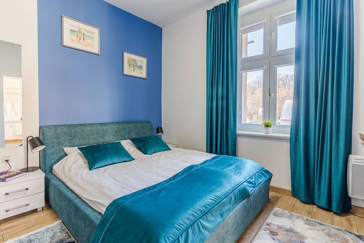 Un dormitorio con cama, mesita de noche, lámpara, ventana con cortinas y dos cuadros en la pared.