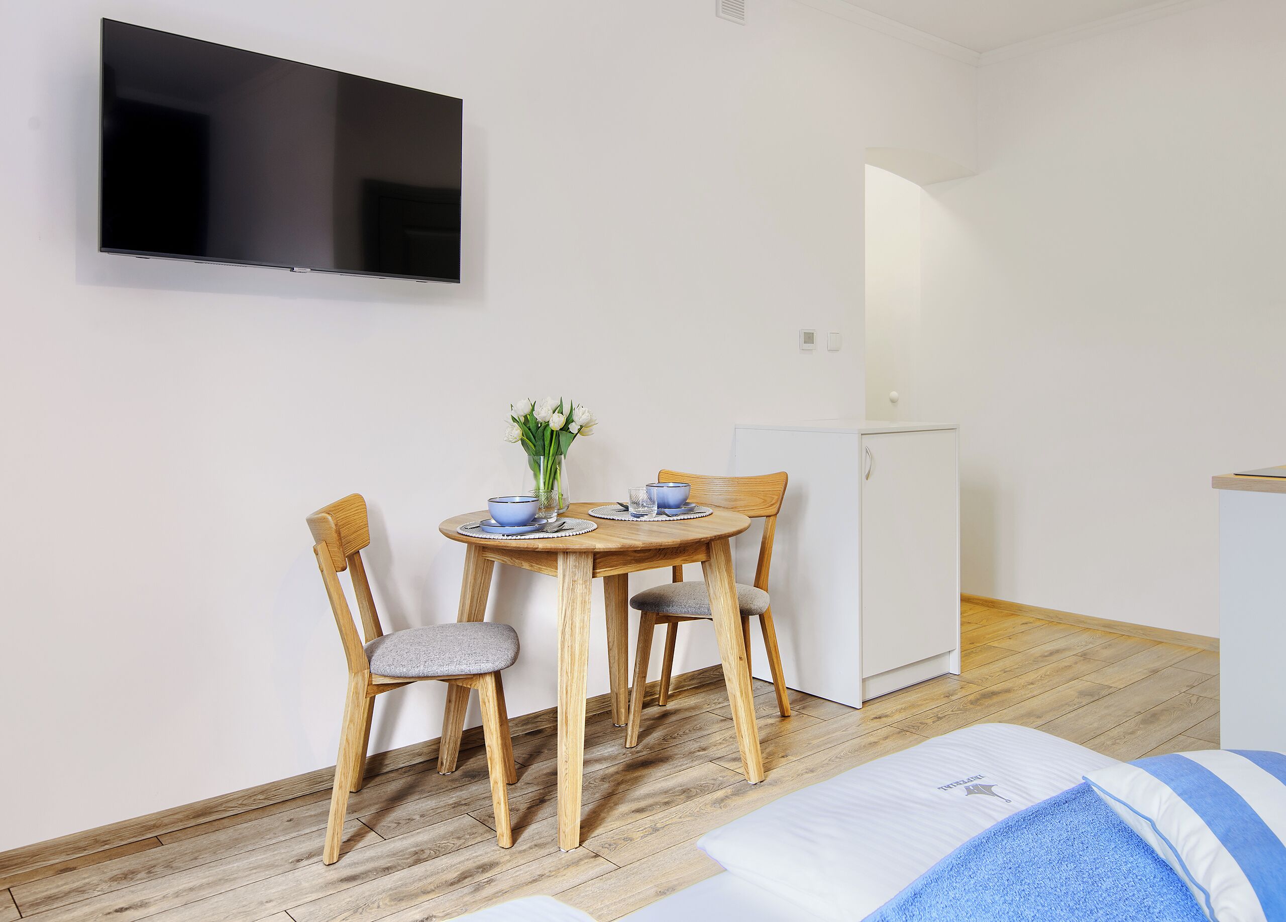 Una habitación pequeña y limpia con una mesa de comedor de madera, dos sillas, un televisor montado en la pared, un refrigerador y una cama con un almohadón azul a rayas.