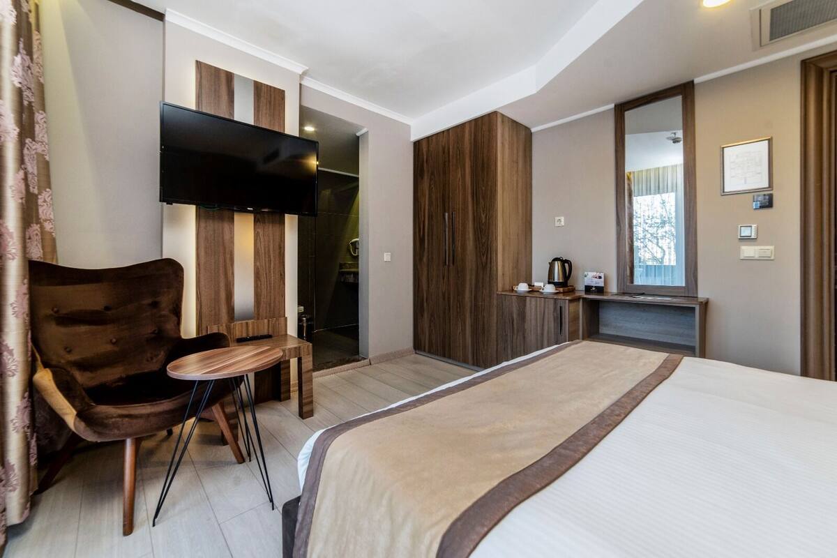 A modern hotel room with a large bed, a wooden chair, a small table, a TV, and a wardrobe.