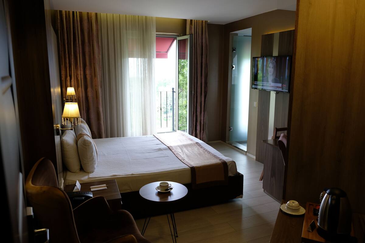 A hotel room with a bed, a desk with a cup and saucer, a chair, a television, and a view of the outdoors.