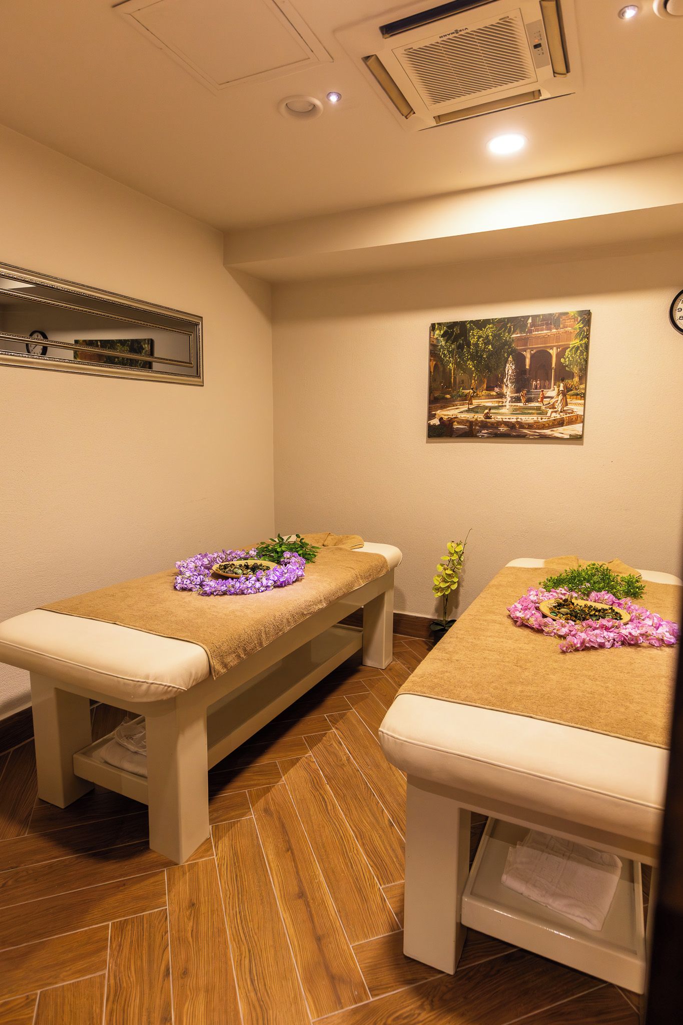 Een ruimte met twee massagetafels, elk voorzien van een handdoek en bloemen. Aan de muur hangt een schilderij en er staat een klok.