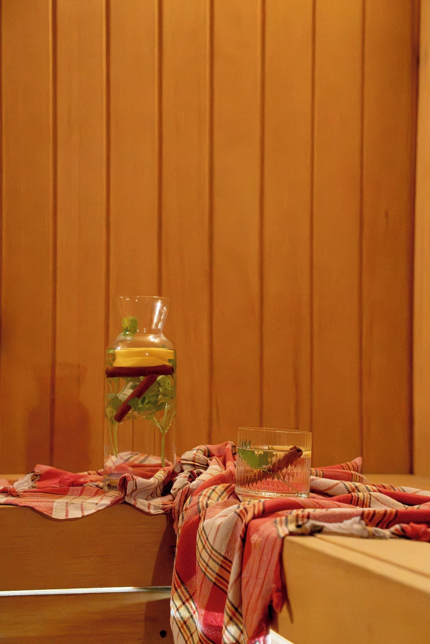 A room with a wooden panel wall, a glass bottle with a plant, and a glass with a plant on a table covered with a plaid cloth.