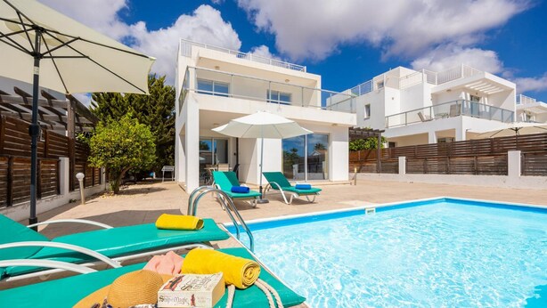 Un área de piscina moderna con sillones de descanso y sombrillas, una piscina azul cristalina y un edificio contemporáneo al fondo.