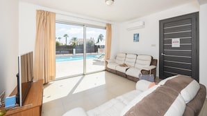 Una habitación moderna con un amplio ventanal corredero que da a una zona de piscina, un sofá, una cama y un televisor sobre un soporte de madera.
