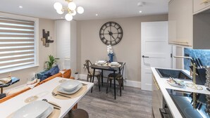 Un moderno comedor con un reloj de pared grande, una mesa de comedor dispuesta con platos y vasos, y una zona de cocina con estufa y utensilios.