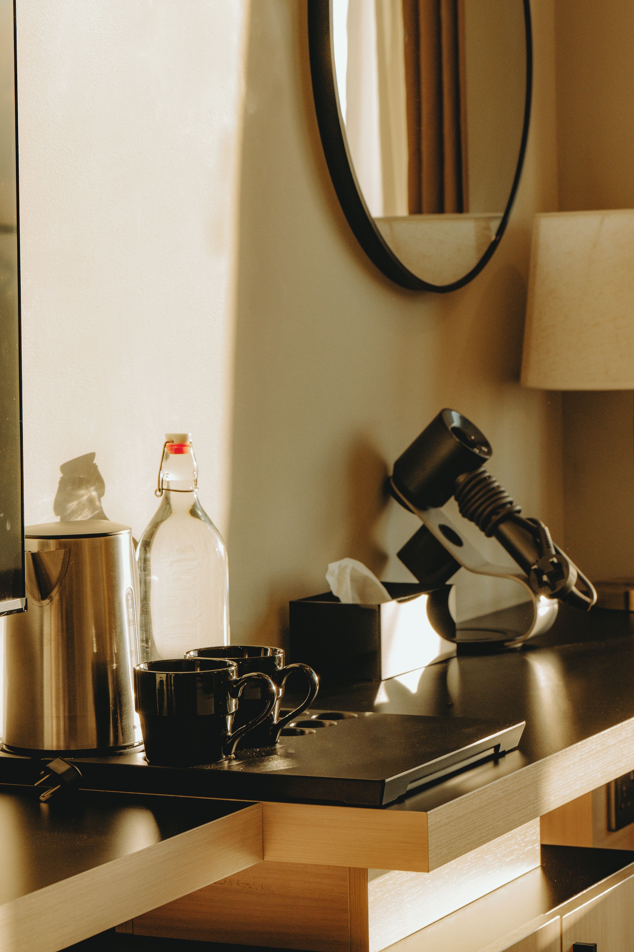 A hotel room with a coffee maker, a bottle, a mug, a hairdryer, and a mirror on the counter.