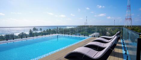 A rooftop pool with lounge chairs, clear skies, and a view of the sea.