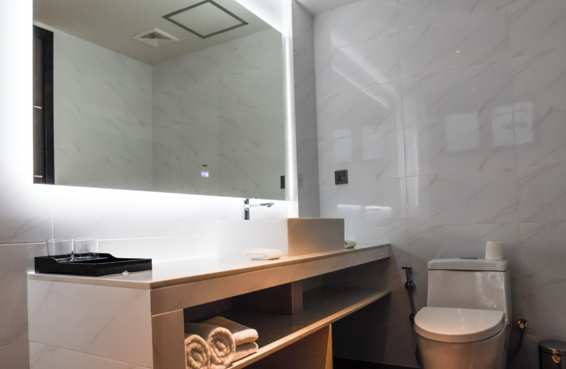 A modern bathroom with a marble wall, a large mirror, a sink with a towel holder, and a toilet.