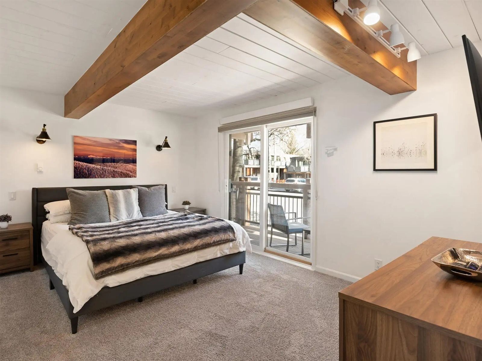 A bedroom with a bed, a wooden ceiling, a balcony with a chair, and a desk with a lamp.