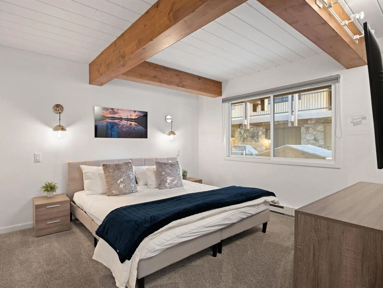 A bedroom with a bed, bedside tables, a nightstand, a window with a view, and a ceiling with exposed beams.