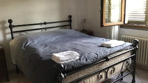 A bed with a dark blue bedspread, a white pillow, and a folded white towel on the footboard. There is a nightstand with a lamp beside the bed. A window with blinds is visible in the background. - Apartment - Sleeps 4 - Pet Friendly (Monsummano Terme)