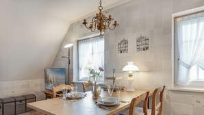 A dining area with a wooden table set for four, surrounded by chairs. A chandelier hangs from the ceiling, and there are two windows with curtains. - Green Teft - thatched house in the southwest part of the village of Keitum (Sylt / OT Keitum)