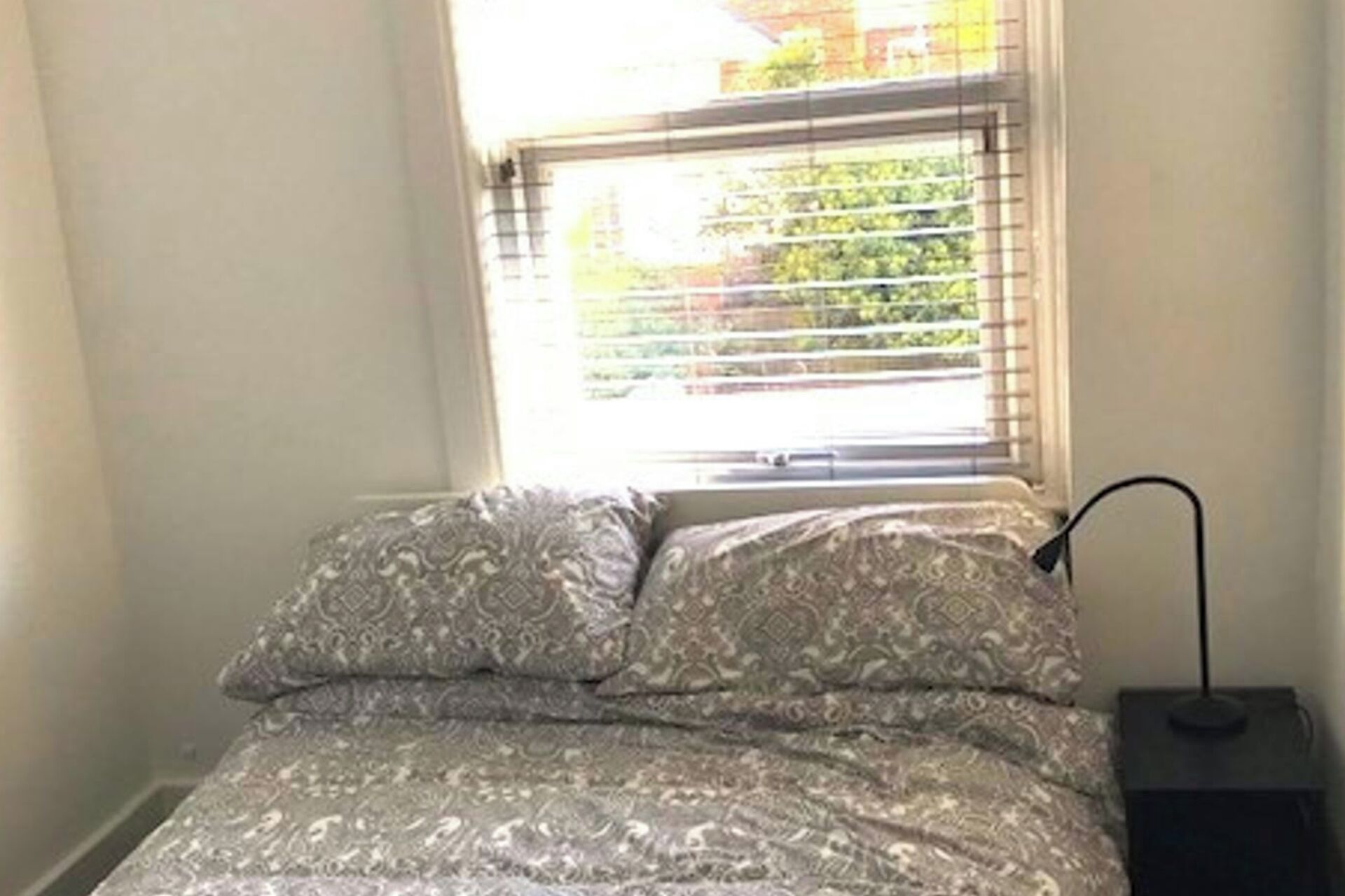 A bedroom with a bed, a window with blinds, and a bedside table with a lamp.
