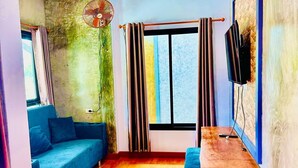 Una sala de estar con un sofá azul, una mesa de centro de madera, un televisor montado en la pared, una ventana con cortinas y un ventilador en la pared.